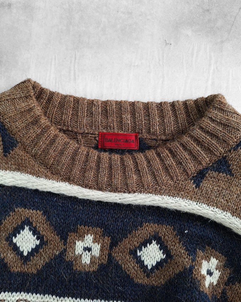 May include: Close-up of a brown, navy, and white patterned sweater. The sweater has a ribbed brown collar and a red tag with the brand name "BALENCIAGA". The design includes geometric shapes and a white stripe.