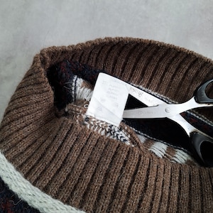 May include: Close-up of a brown knit sweater collar with a striped pattern in white, navy, and burgundy. Silver scissors with black handles are cutting a white tag attached to the sweater.