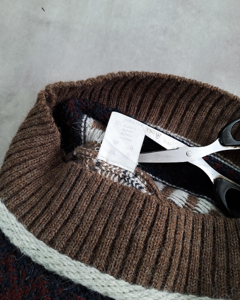 May include: Close-up of a brown knit sweater collar with a striped pattern in white, navy, and burgundy. Silver scissors with black handles are cutting a white tag attached to the sweater.