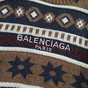 May include: Close-up of a brown, navy, and white patterned jumper. The jumper features a repeating geometric design and the text "BALENCIAGA PARIS" in white and red. The jumper has a ribbed neckline.