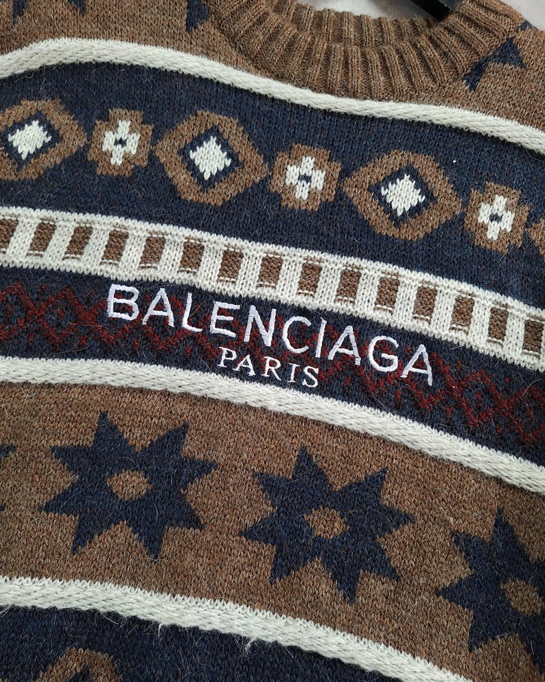 May include: Close-up of a brown, navy, and white patterned jumper. The jumper features a repeating geometric design and the text "BALENCIAGA PARIS" in white and red. The jumper has a ribbed neckline.