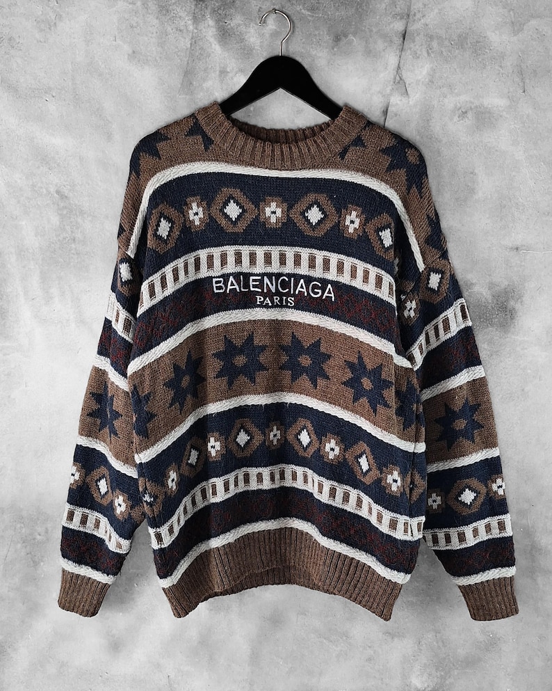 May include: A patterned sweater in shades of brown, navy, and white, with the text "BALENCIAGA PARIS". The sweater has a crew neck and long sleeves. The design incorporates geometric shapes and star motifs.