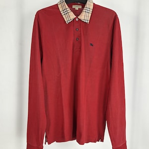 Puede incluir: Un polo rojo de manga larga con cuello de cuadros en beige, negro y rojo. La prenda cuenta con una tapeta de tres botones y un pequeño logotipo oscuro bordado en el pecho izquierdo. Se muestra colgado en una percha de color negro.