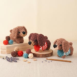 May include: Three handmade crochet dachshund dogs in shades of brown, each with a unique sweater. One wears a red and white patterned sweater, another a blue and white one. Accessories include yarn balls and wooden buttons.
