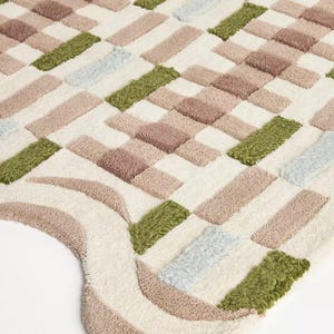 Peut inclure: Gros plan sur un tapis doux et textur&eacute; avec un motif g&eacute;om&eacute;trique. Le tapis pr&eacute;sente des blocs rectangulaires dans des tons cr&egrave;me, marron, vert et bleu clair. Le bord du tapis a un design incurv&eacute; avec des rayures cr&egrave;me et marron.