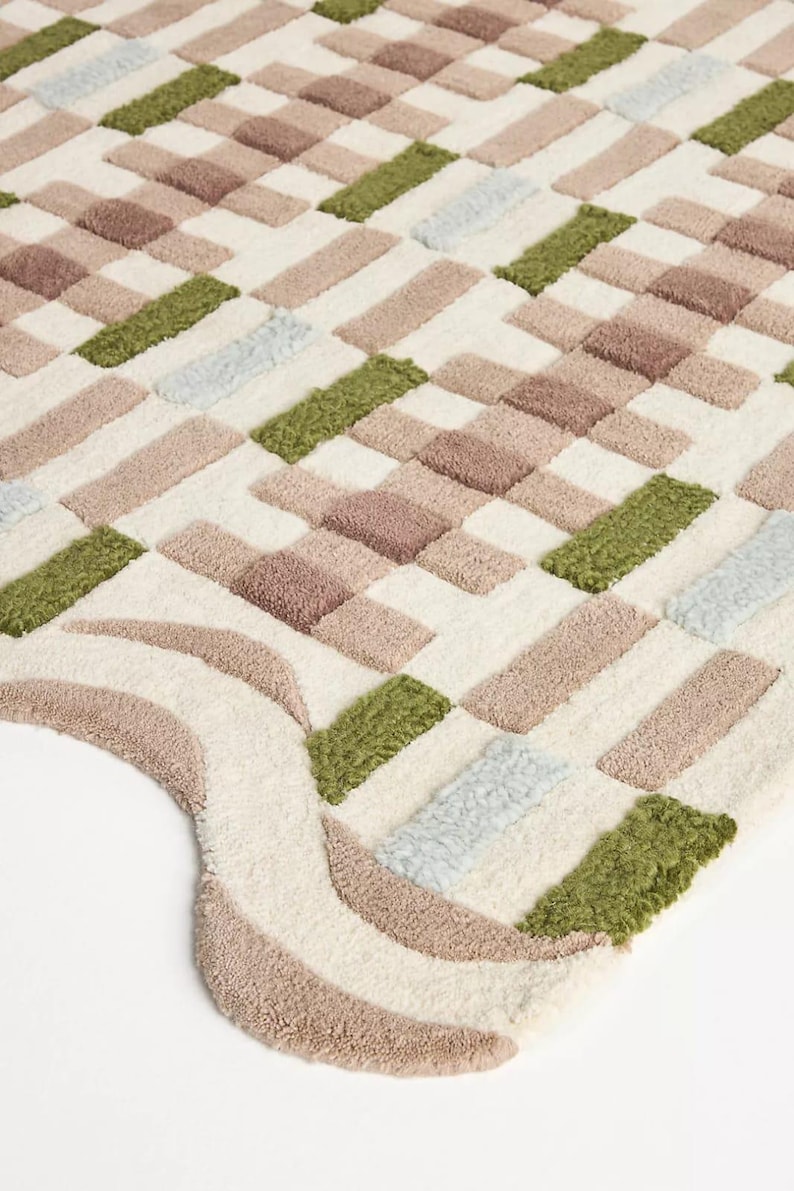 Peut inclure: Gros plan sur un tapis doux et textur&eacute; avec un motif g&eacute;om&eacute;trique. Le tapis pr&eacute;sente des blocs rectangulaires dans des tons cr&egrave;me, marron, vert et bleu clair. Le bord du tapis a un design incurv&eacute; avec des rayures cr&egrave;me et marron.