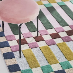 May include: A colorful geometric rug with a checkerboard and striped pattern in shades of pink, green, yellow, and blue. A pink velvet chair with black metal legs is in the foreground. The rug is a decorative home furnishing.