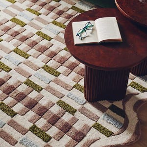 Peut inclure: Tapis &agrave; motifs g&eacute;om&eacute;triques dans les tons beige, marron, vert et bleu. Une petite table ronde marron fonc&eacute; avec un livre ouvert et des lunettes. Le bord du tapis a une bordure ondul&eacute;e et textur&eacute;e.