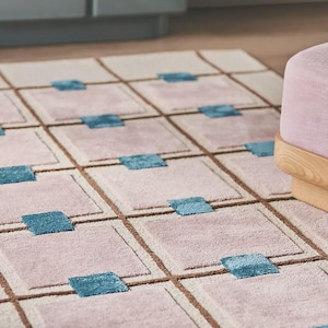 Pode incluir: Um tapete retangular com um padrão geométrico. O tapete apresenta uma grade de quadrados em rosa claro e bege, com quadrados de destaque azul-petróleo. Linhas marrons definem a grade. O tapete está em um piso de madeira.