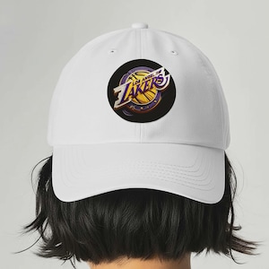 May include: White baseball cap featuring the Los Angeles Lakers logo. The logo is a circular design with the team name and a basketball graphic in purple, gold, and black. The cap is a classic six-panel design.
