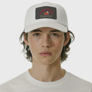 Könnte beinhalten: Weißes Baseball-Cap mit gebogenem Schirm. Auf dem Cap befindet sich ein rechteckiger Aufnäher mit den Worten "Happy Fields Forever" in Rot und Weiß, verziert mit floralen Elementen. Das Cap ist aus weichem Material.