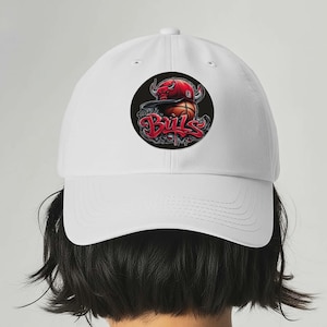 Puede incluir: Gorra de béisbol blanca con un diseño gráfico circular que muestra una pelota de baloncesto, una cabeza de toro y la palabra "Bulls" en rojo. La gorra tiene una visera curva y una corona estructurada.