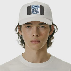 Op de afbeelding: Witte baseballpet met een rechthoekige patch met het woord "ICEMAN" boven een blauw-witte afbeelding van een vogel. De pet heeft een gebogen klep en een donkergrijze patch.