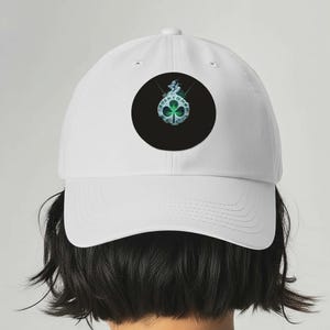 Könnte beinhalten: Weiße Baseballkappe mit einem schwarzen runden Aufnäher mit einem grünen Kleeblatt-Design. Der Aufnäher enthält auch den Text "BOSTON" in kreisförmiger Anordnung. Die Kappe ist aus glattem Stoff gefertigt und hat einen gebogenen Schirm.
