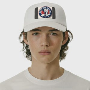 Könnte beinhalten: Eine weiße Baseballkappe mit einem rechteckigen Aufnäher. Der Aufnäher zeigt ein kreisförmiges Design mit einem Elefanten, Sternen und dem Text "REDRAWN & READY". Das Design ist von einem schwarzen Rand umgeben.