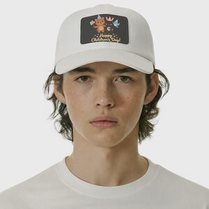 May include: White baseball cap with a rectangular patch featuring a teddy bear, flowers, and the text "Happy Children's Day!". The patch has a dark background with a crown and a blue bird. The cap is worn by a person.