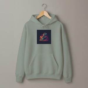 May include: A sage green hoodie with a kangaroo pocket and a drawstring hood. The front features a square graphic of a cartoon turtle with a blue mask and colorful accents. The hoodie is hanging on a wooden hanger.