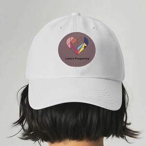 May include: White baseball cap featuring a circular patch with a geometric heart design in pink, brown, yellow, blue, and green. The text "Love's Frequency" is printed below the heart. The cap has a curved brim and a button on top.