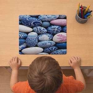 Puede incluir: Un rompecabezas cuadrado que presenta una colección de piedras ovaladas decoradas con varios patrones azules y blancos, junto con algunas piedras rosas y blancas, dispuestas en un grupo denso sobre una superficie de madera clara junto a un portalápices.