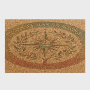 Può includere: Uno zerbino rettangolare in fibra di cocco color cuoio con un disegno centrale a rosa dei venti circondato da una corona di rami d'ulivo. Un bordo ovale incornicia la grafica, contenente il testo HUMANITY HAS NO BORDERS in lettere maiuscole verde scuro.