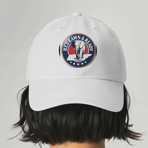 Op de afbeelding: Witte baseballcap met een cirkelvormig embleem. Het embleem toont de tekst "REDRAWN & READY" rond een blauw en rood ontwerp, met een olifant en sterren.