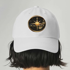 May include: White baseball cap featuring a gold and black compass design with the text "THE DEALMAKER." The cap has a curved brim and a classic six-panel construction. The design includes a map of the world.