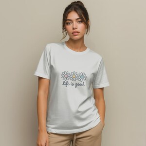 Può includere: Una t-shirt bianca a maniche corte con una grafica di tre margherite con centri color pastello rosa, giallo e blu. Sotto i fiori, il testo "life is good" è stampato in un carattere corsivo grigio scuro sulla zona del petto.