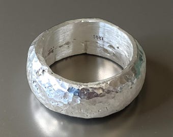 May include: A hammered silver ring with a textured surface. The ring has a wide band and a slightly irregular shape. The metal reflects light, creating highlights and shadows. The ring appears to be handmade.