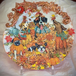 Könnte beinhalten: Dekorative Keramikplatte mit einer Herbstszene mit mehreren Hunden in einem Haufen von Herbstblättern. Die Hunde tragen Pullover und Kostüme, mit Kürbissen und Herbstlaub. Die Platte hat einen dekorativen Goldrand.