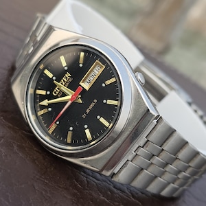 Pode incluir: Um relógio de pulso automático Citizen vintage com mostrador preto, marcadores dourados e ponteiro de segundos vermelho. O relógio exibe o dia e a data e possui uma pulseira de aço inoxidável prateado. Um relógio clássico.