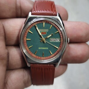 May include: A vintage Seiko 5 automatic watch with a green dial and a red and gold bezel. The watch has a brown leather strap. The day and date are displayed at the 3 o'clock position. The text "SEIKO 5 AUTOMATIC" is visible on the watch face.