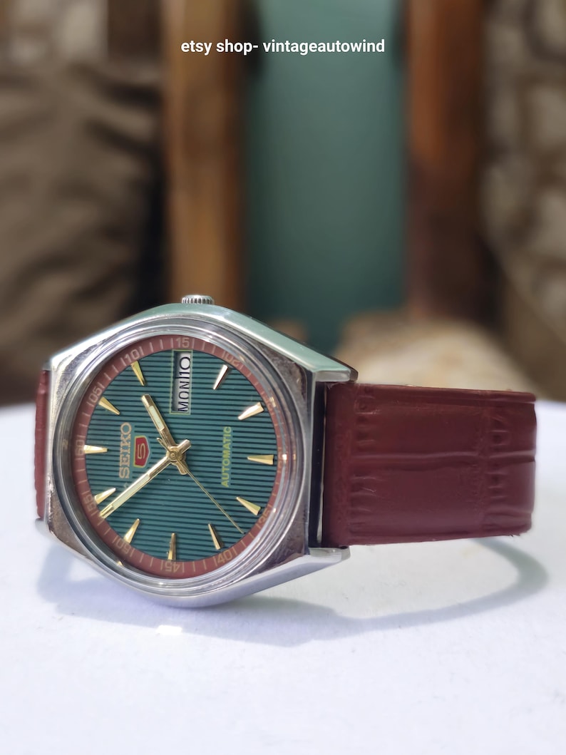 May include: A vintage Seiko automatic watch with a teal face, gold hands and markers, and a burgundy leather strap. The watch has a silver-tone case and the text "SEIKO" and "AUTOMATIC" are visible on the face. The strap is approximately 0.75 inches wide.