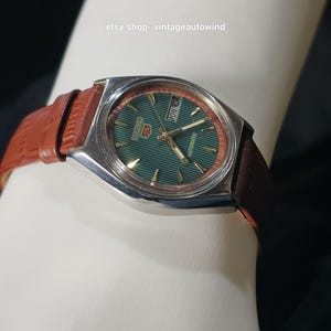 May include: A vintage Seiko 5 automatic watch with a green dial and a brown leather strap. The watch has a silver-colored case and a day-date display. The strap is a reddish-brown color.