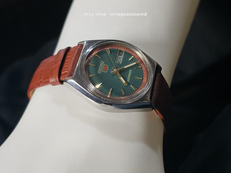 May include: A vintage Seiko 5 automatic watch with a green dial and a brown leather strap. The watch has a silver-colored case and a day-date display. The strap is a reddish-brown color.