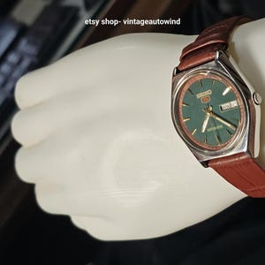 May include: A vintage Seiko watch with a green dial and a brown leather strap. The watch has a silver-colored case and displays the day and date. The watch is on a white surface.