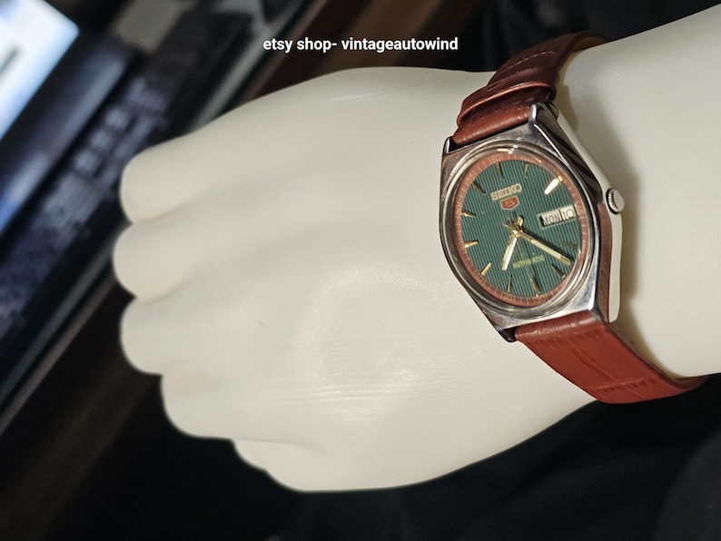 May include: A vintage Seiko watch with a green dial and a brown leather strap. The watch has a silver-colored case and displays the day and date. The watch is on a white surface.