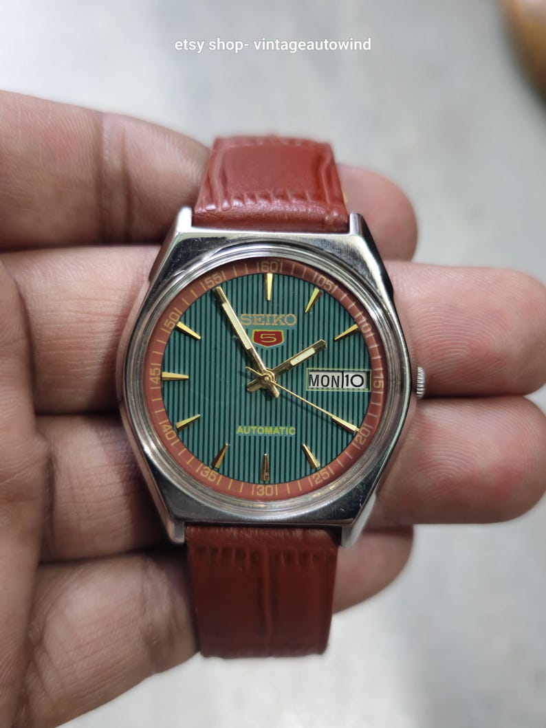 May include: A vintage Seiko 5 automatic watch with a green dial and a red and gold bezel. The watch has a brown leather strap. The day and date are displayed at the 3 o'clock position. The text "SEIKO 5 AUTOMATIC" is visible on the watch face.