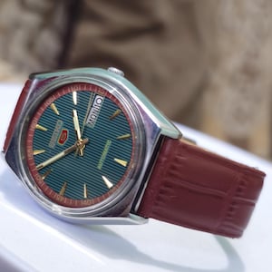 May include: A vintage Seiko watch with a teal dial and gold accents. The watch features a red and silver bezel, a brown leather strap, and the day of the week displayed. The watch face has a striped pattern.
