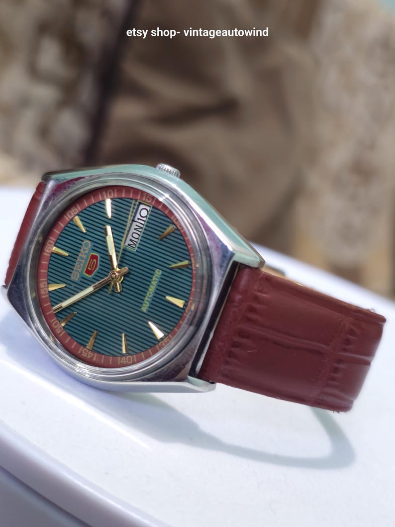 May include: A vintage Seiko watch with a teal dial and gold accents. The watch features a red and silver bezel, a brown leather strap, and the day of the week displayed. The watch face has a striped pattern.