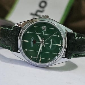 May include: A silver-toned Seiko automatic wristwatch with a green face and a date window. The watch has a green leather band with green stitching. The watch face has white hands and markers.
