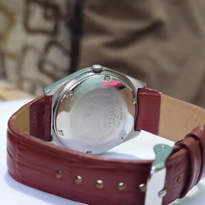 May include: A vintage Seiko watch with a silver-toned case and a burgundy leather strap. The watch face is not visible, but the back of the watch shows the Seiko brand and model details. The strap is secured with a silver buckle.