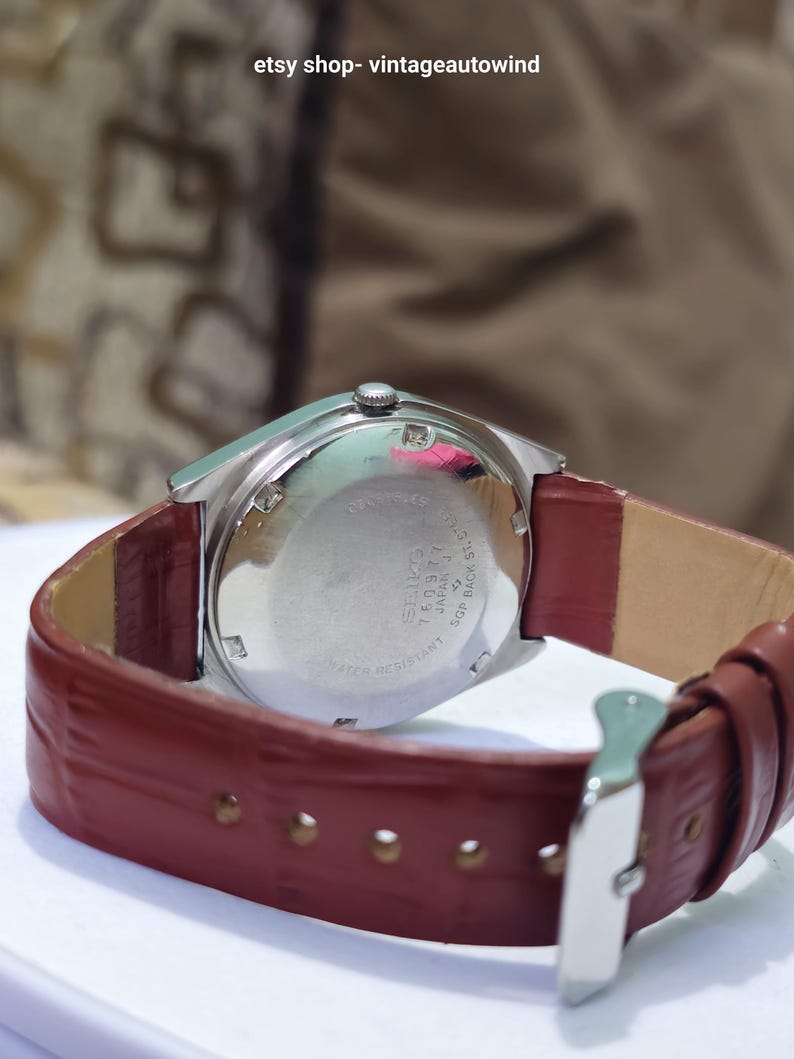 May include: A vintage Seiko watch with a silver-toned case and a burgundy leather strap. The watch face is not visible, but the back of the watch shows the Seiko brand and model details. The strap is secured with a silver buckle.
