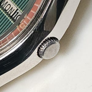 May include: Close-up of a vintage wristwatch with a silver-toned case and a green dial. The dial displays the day and date, with "MON 16" visible. The watch has a textured crown and a brick-patterned inner ring. The Etsy shop name "vintageautowind" is visible.