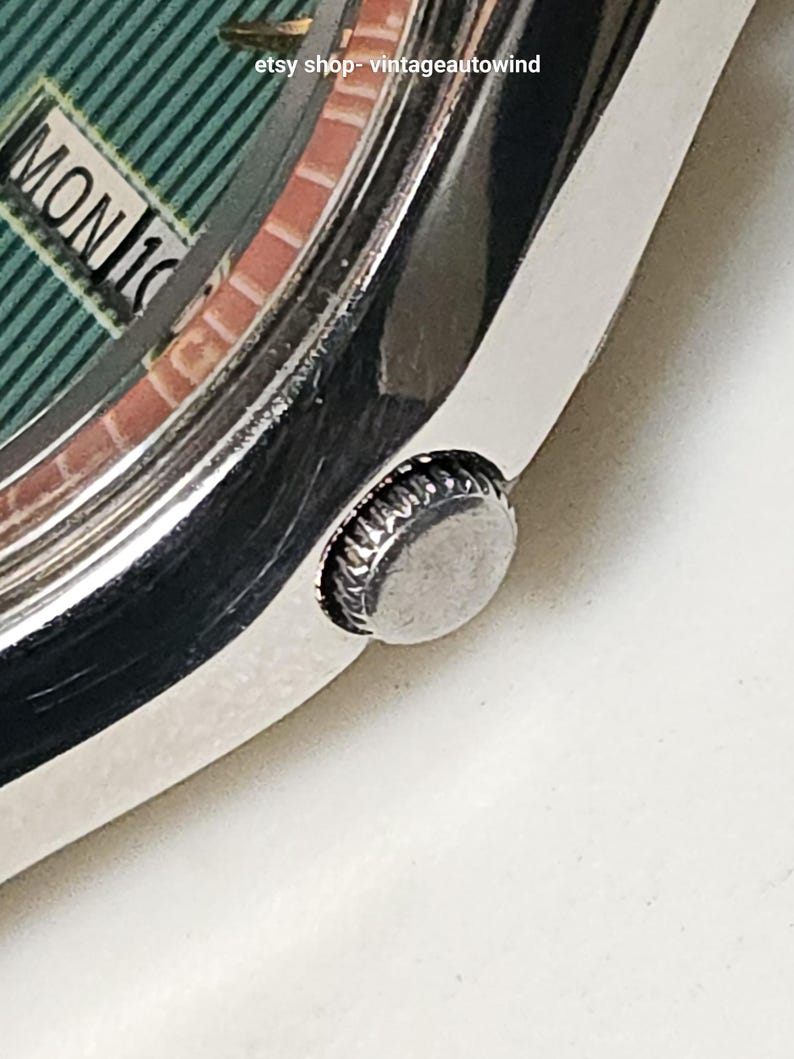May include: Close-up of a vintage wristwatch with a silver-toned case and a green dial. The dial displays the day and date, with "MON 16" visible. The watch has a textured crown and a brick-patterned inner ring. The Etsy shop name "vintageautowind" is visible.