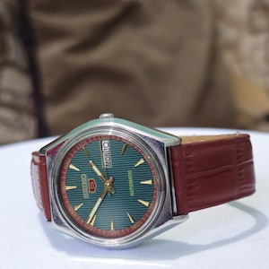 May include: A vintage Seiko 5 automatic watch with a green dial and gold accents. The watch has a silver-tone case and a brown leather strap. The day of the week is displayed at the top. The watch is a classic timepiece.