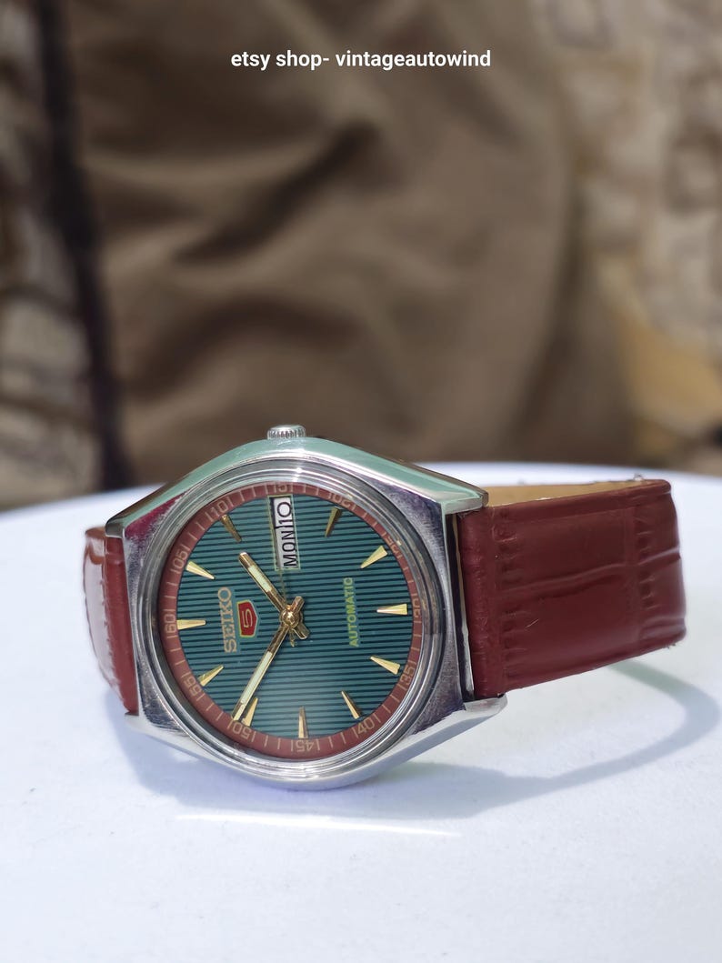 May include: A vintage Seiko 5 automatic watch with a green dial and gold accents. The watch has a silver-tone case and a brown leather strap. The day of the week is displayed at the top. The watch is a classic timepiece.