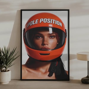 Puede incluir: Impresión enmarcada que presenta a una mujer con un casco de carreras naranja brillante. El casco tiene las palabras "POLE POSITION" en blanco. La imagen está sobre un fondo neutro, con una planta y una pequeña lámpara en primer plano.