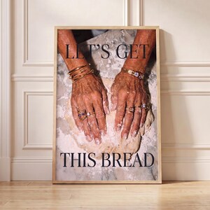 Op de afbeelding: Een ingelijste verticale wandkunstprint met twee handen bedekt met bloem die een deegbal kneden. De handen zijn versierd met meerdere gouden armbanden en ringen. De tekst "LET'S GET" staat bovenaan en "THIS BREAD" onderaan.