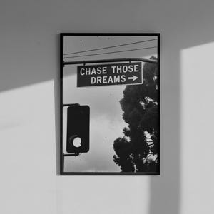 Może przedstawiać: Czarno-biała oprawiona grafika przedstawiająca scenę uliczną ze światłami drogowymi i znakiem z napisem "CHASE THOSE DREAMS" ze strzałką. W tle widoczne jest drzewo. Grafika jest zamontowana na białej ścianie.