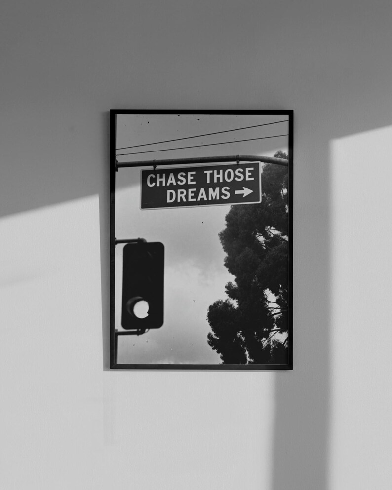 Może przedstawiać: Czarno-biała oprawiona grafika przedstawiająca scenę uliczną ze światłami drogowymi i znakiem z napisem "CHASE THOSE DREAMS" ze strzałką. W tle widoczne jest drzewo. Grafika jest zamontowana na białej ścianie.